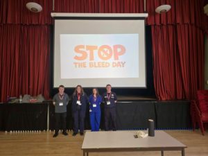 Four individuals stood in front of a stage, with a presentation titles "Stop the Bleed Day"