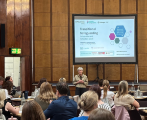 Participants attend a Transitional Safeguarding event in a large wood-panelled conference hall. THe Deputy Mayor for Safer and Stronger communities stands at the front of the room speaking to attendees seated at round tables. Behind her, a presentation slide displaying "Transitional Safeguarding" is projected onto a large screen. Attendees are listening, taking notes and engaging in discussion as part of the showcase event focused on improving support for young people experiencing complex safeguarding risks.