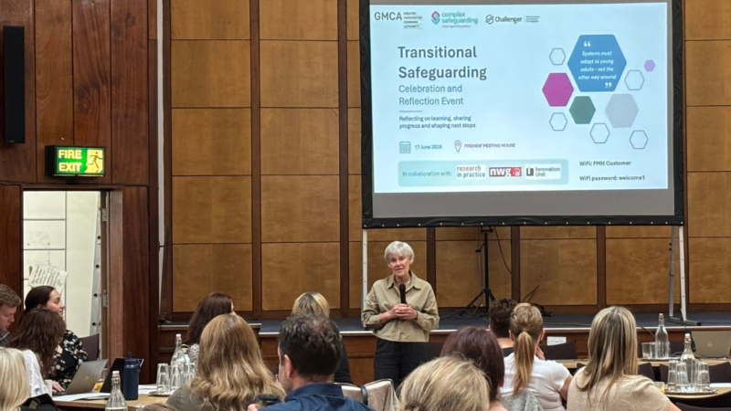 Participants attend a Transitional Safeguarding event in a large wood-panelled conference hall. THe Deputy Mayor for Safer and Stronger communities stands at the front of the room speaking to attendees seated at round tables. Behind her, a presentation slide displaying "Transitional Safeguarding" is projected onto a large screen. Attendees are listening, taking notes and engaging in discussion as part of the showcase event focused on improving support for young people experiencing complex safeguarding risks.