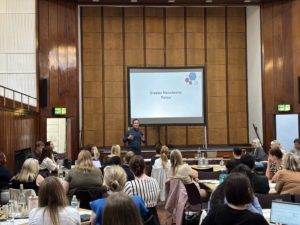 Participants attend a Transitional Safeguarding event in a large wood-panelled conference hall. A presenter from Greater Manchester Police stands at the front of the room speaking to attendees seated at round tables. Behind him, a presentation slide displaying "Greater Manchester Police" is projected onto a large screen. Attendees are listening, taking notes and engaging in discussion as part of the showcase event focused on improving support for young people experiencing complex safeguarding risks.